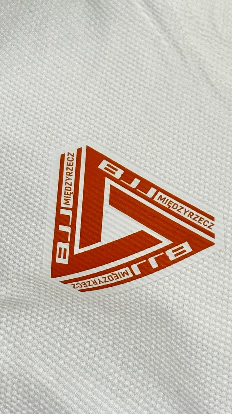 Pomarańczowe trójkątne logo BJJ Międzyrzecz na białym kimonie