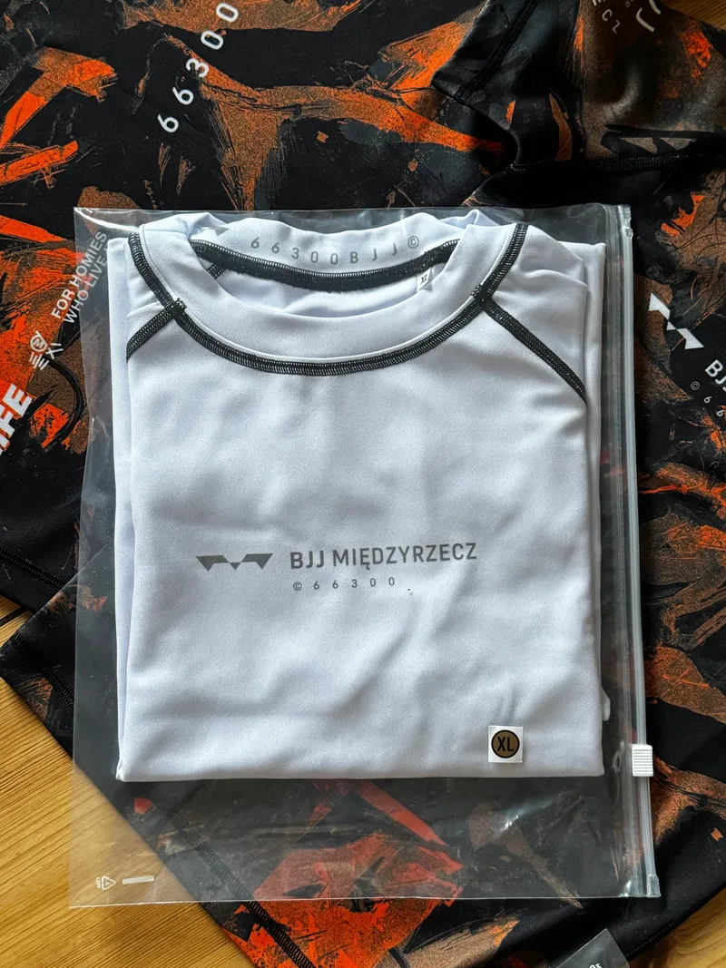 Biały rashguard BJJ Międzyrzecz w opakowaniu na tle pomarańczowych rashguardów