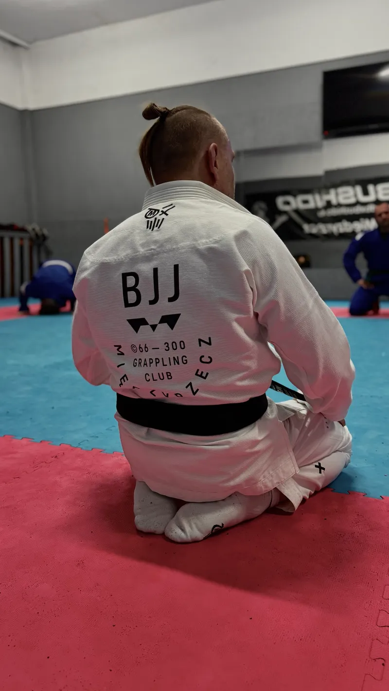 Zawodnik w białym kimonie z logo klubu na plecach siedzi na macie